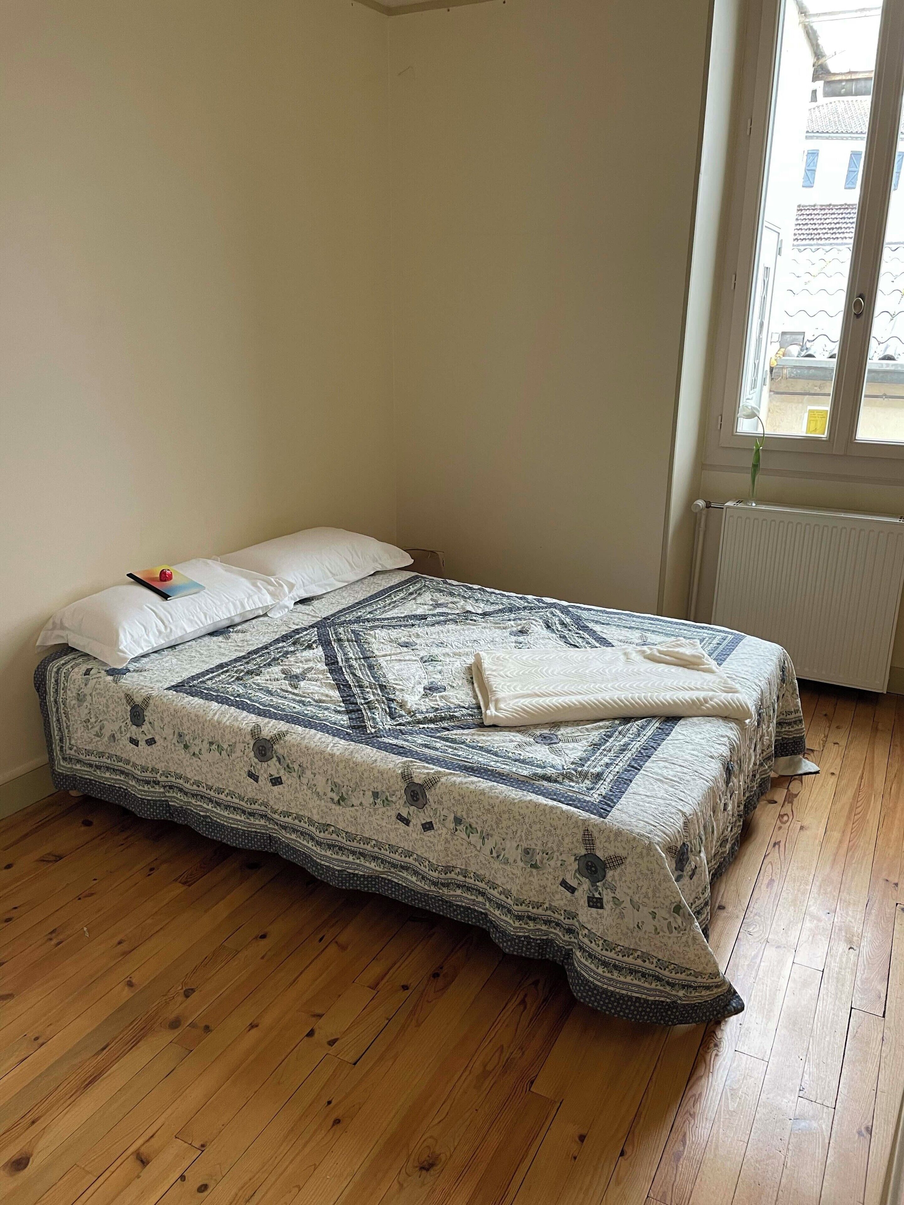 A single bed with a patterned bedspread and pillows, a radiator, and a window with a view of buildings outside.