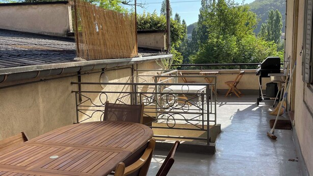 Een terras op het dak met een houten tafel en stoelen, een barbecue en uitzicht op groen en heuvels.