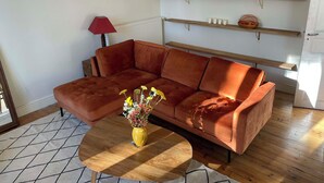 Een woonkamer met een bruine hoekbank, een gele vaas met bloemen, een ronde houten salontafel en houten planken boven de bank.