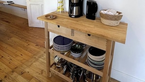 Een keukenhoek met een houten tafel waarop een lamp, koffiezetapparaat en mandje staan. Onder de tafel bevinden zich planken met servies en bestek.