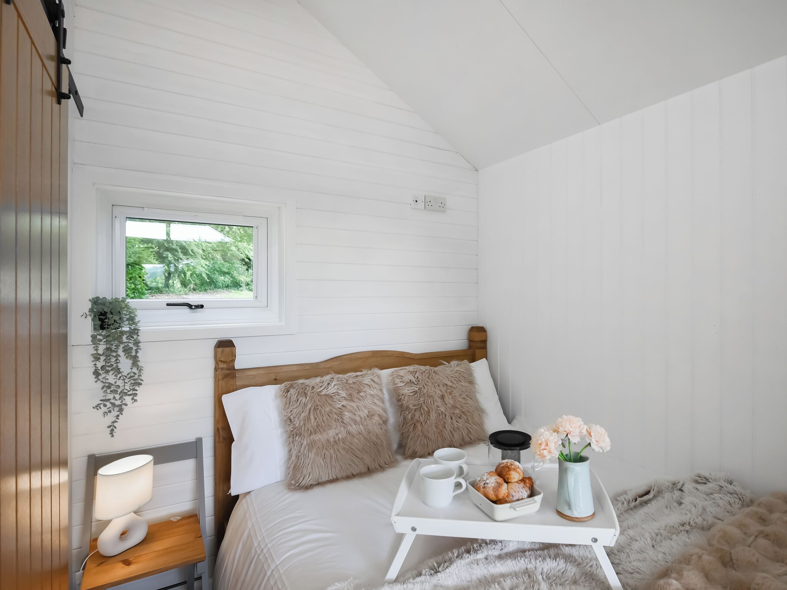 Une chambre avec un lit en bois, une table de chevet avec une lampe, une fenêtre donnant sur un paysage verdoyant et un plateau avec des pâtisseries et des fleurs.