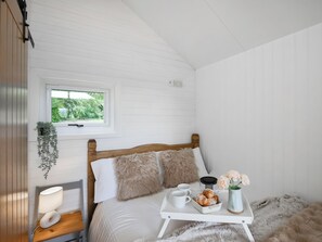 A bedroom with a wooden bed, a bedside table with a lamp, a window with a view of greenery, and a tray with pastries and flowers.