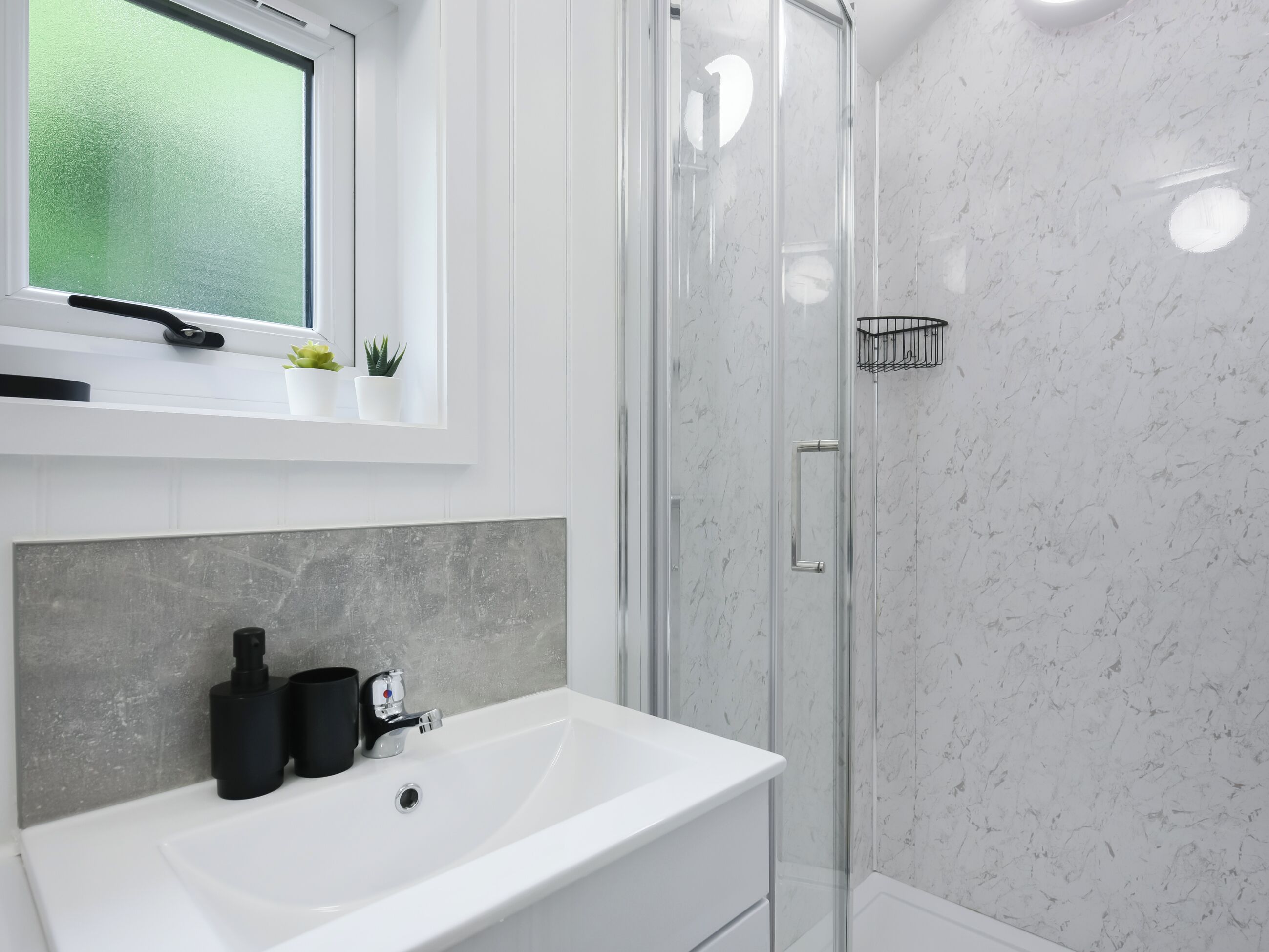 A modern bathroom with a white sink, a glass shower enclosure, and a window with a frosted glass view.