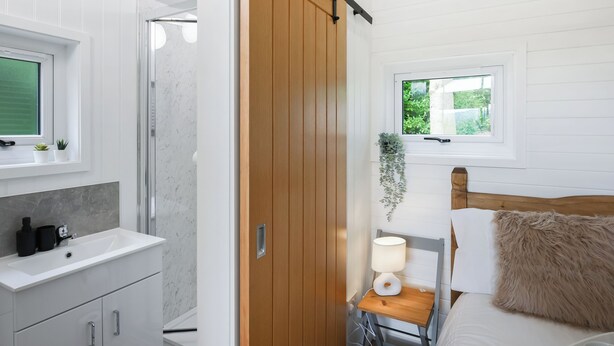 Un baño moderno con un tocador blanco, una puerta de madera, una mesita de noche con lámpara y una ventana con vistas a la vegetación.