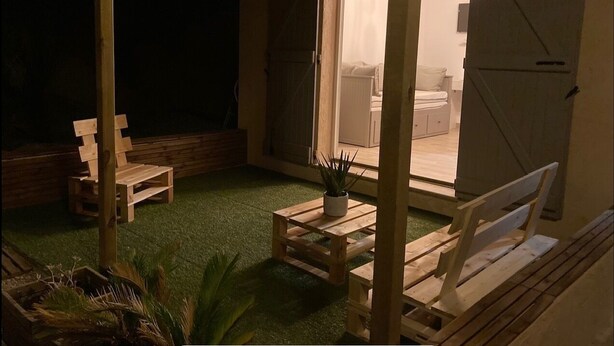Een terras met houten banken en een kleine tafel met een plant, omgeven door een houten hek en met uitzicht op de binnenruimte.