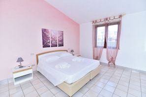 A neatly made bed with white linens and towels, a bedside table with a lamp, and a window with sheer curtains.