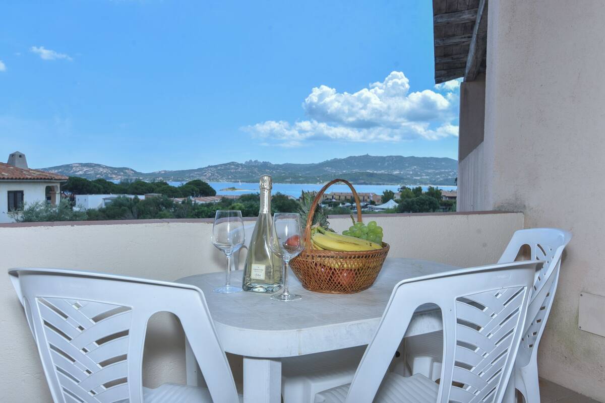 Ein Tisch mit einer Flasche und Gläsern, ein Obstkorb und weiße Plastikstühle auf einem Balkon mit Blick auf eine schöne Aussicht.