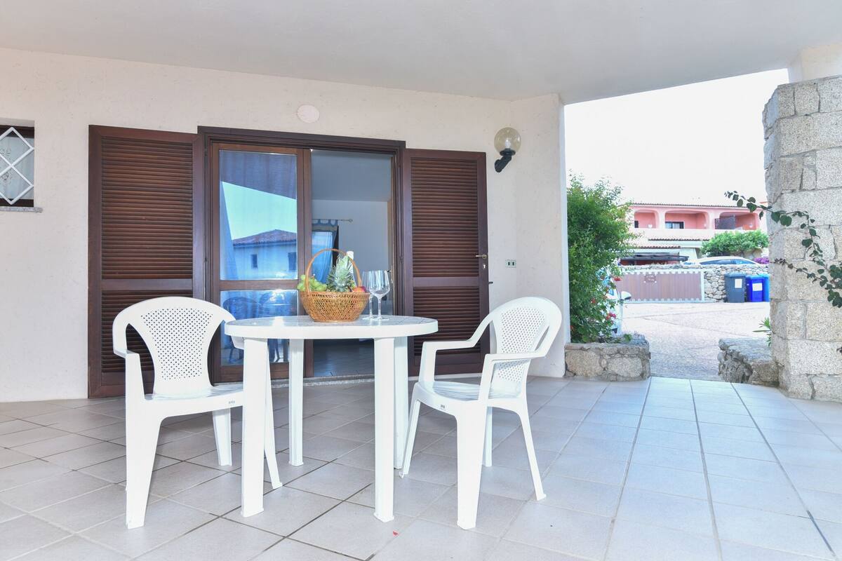 Eine Terrasse mit Tisch und zwei weißen Stühlen, ein Korb voller Früchte und einem Glas, sowie ein Blick auf ein Gebäude im Hintergrund.