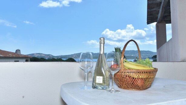 A table with a bottle of white wine, two glasses, and a wicker basket containing fruits on a balcony with a clear view of the sky and distant mountains. - La Costa Mare (Arzachena)