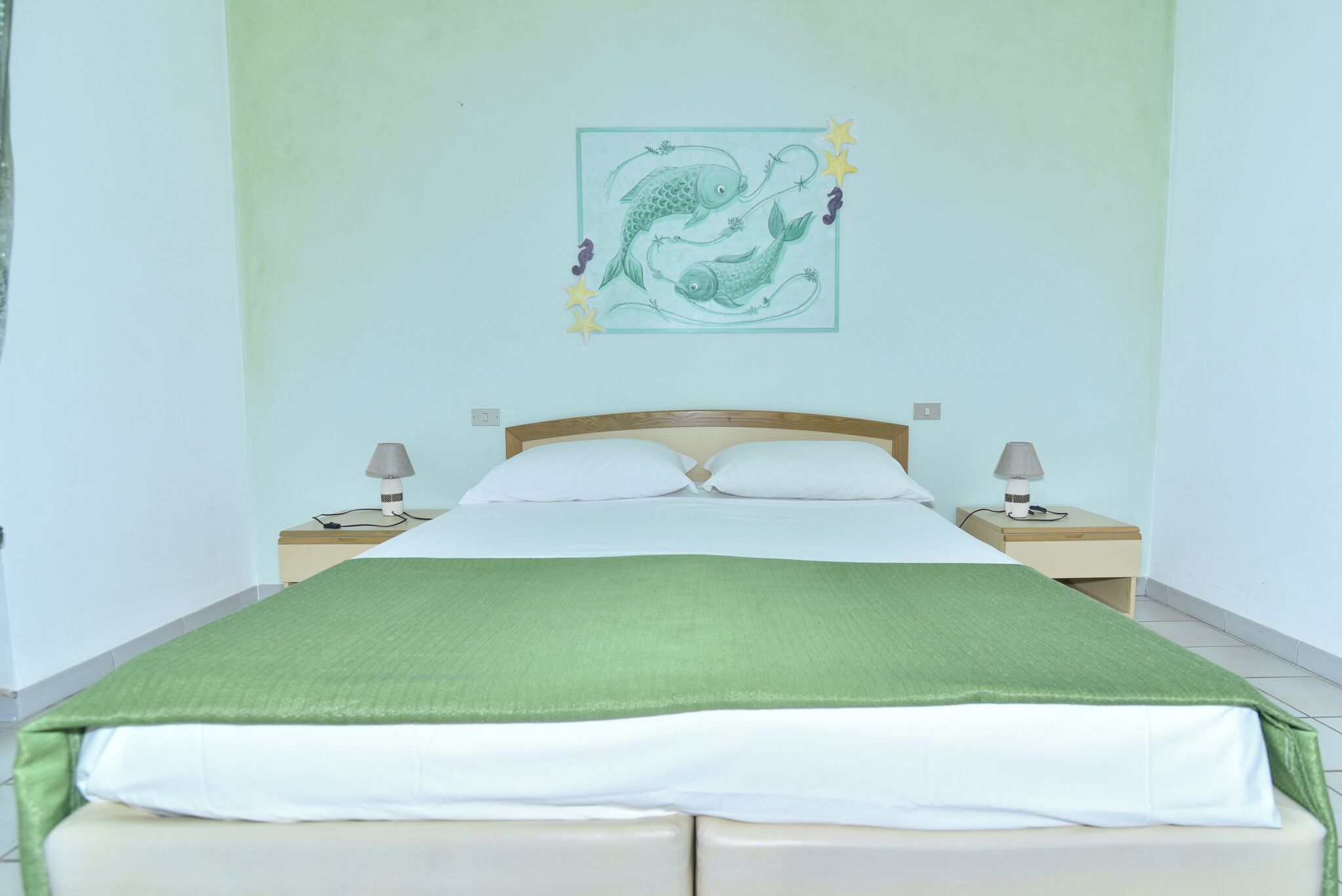 Ein ordentlich bezogenes Bett mit grüner Decke, zwei Nachttischlampen und einem Wandbild von Fischen.