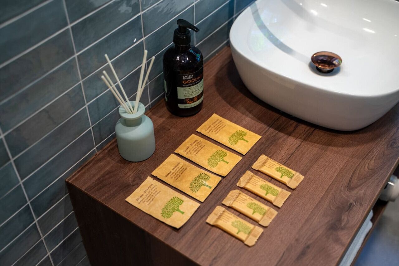 A bathroom with a wooden countertop, a white sink, a bottle of soap, a vase with reed diffusers, and several small brown packets.