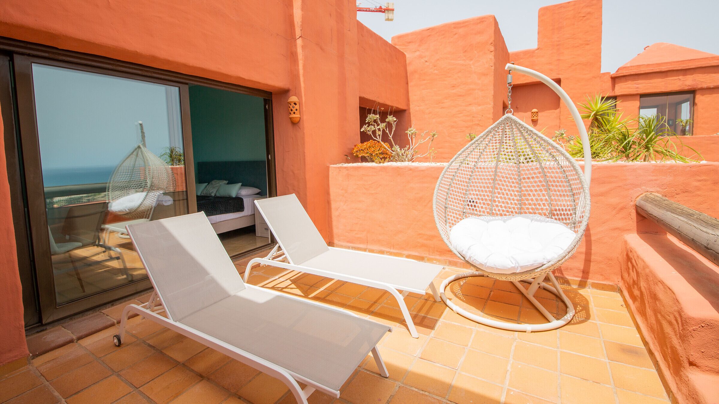 A balcony with two white lounge chairs, a hanging egg chair, and a glass door leading to a bedroom.