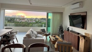 A modern living room with a large window offering a city view, a flat-screen TV mounted on the wall, and a dining area with a wooden table and chairs. - Luxury Retreat, VGolf. Marina Vallarta. (Puerto Vallarta)