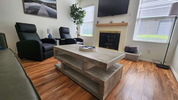 Un salon avec une table basse en bois, des fauteuils en cuir noir, une cheminée, un téléviseur et une grande fenêtre avec des stores.