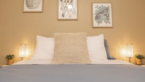 A neatly made bed with white linens and a textured pillow, flanked by two bedside lamps with plants. Three framed artworks are hung on the wall above the bed. - Adventurers Base Camp! (Boise)