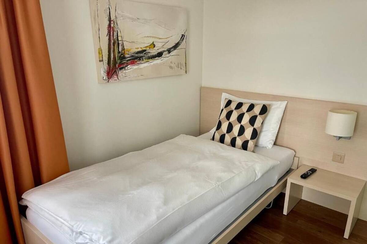 A single bed with a white mattress and a headboard with a patterned pillow. There is a bedside table with a lamp and a remote control on it.
