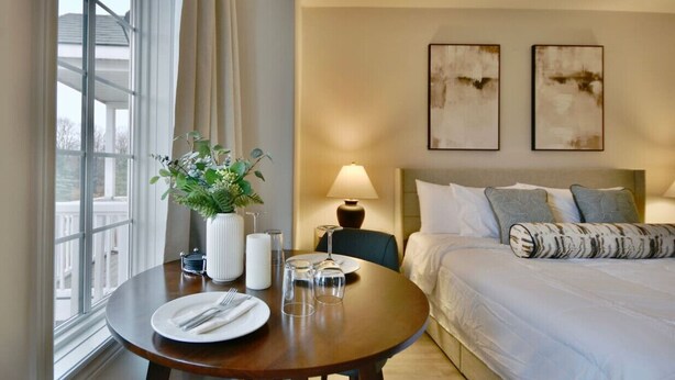 A hotel room with a bed, a bedside table with a lamp, a window with curtains, and two abstract paintings on the wall.