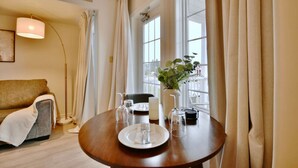 A dining area with a round table set for two, a sofa, a floor lamp, and a window with curtains.