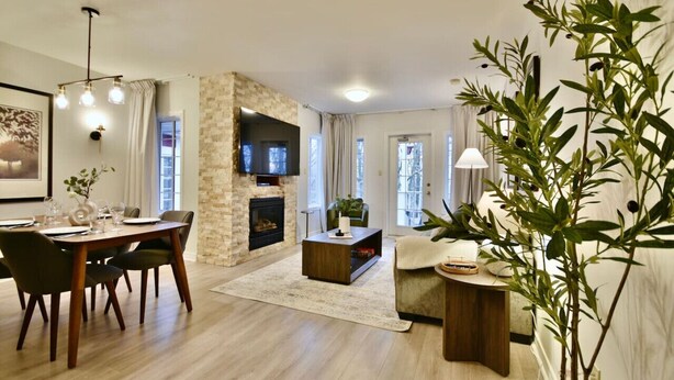 Een ruime woonkamer met een stenen openhaard, een eethoek met een tafel en stoelen, en een gezellige zithoek met een bank en een lamp.