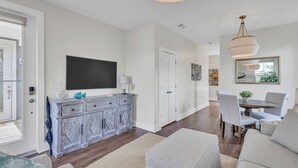 A spacious living room with a large flat-screen TV, a wooden cabinet, and a dining area with a round table and chairs.