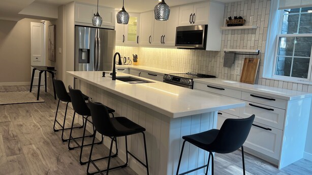 A modern kitchen with a large island, black bar stools, stainless steel appliances, pendant lighting, and a stone backsplash.