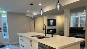 A modern kitchen with a central island, stainless steel appliances, pendant lighting, and a flat-screen TV mounted on the wall.