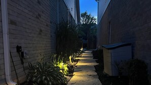 A narrow pathway flanked by buildings with a brick exterior and a small garden area with plants and a pathway light.
