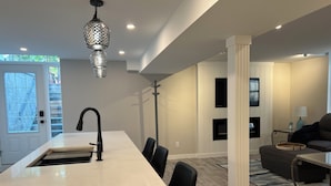 A modern kitchen with a white countertop and black chairs, a pendant light, and a dining area with a sofa and a coffee table.
