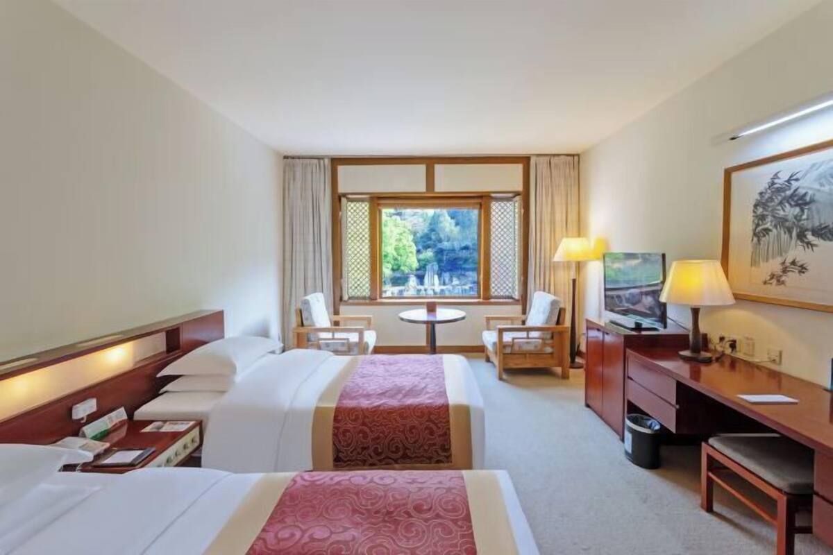 A hotel room with two beds, a desk, a chair, a small table, and a window with a view.