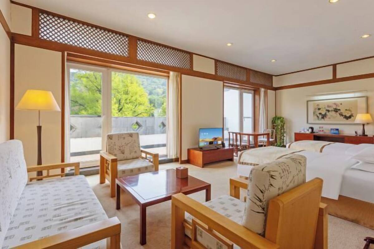 A hotel room with a sofa, chairs, a TV, a bed, and a balcony with a view of trees.