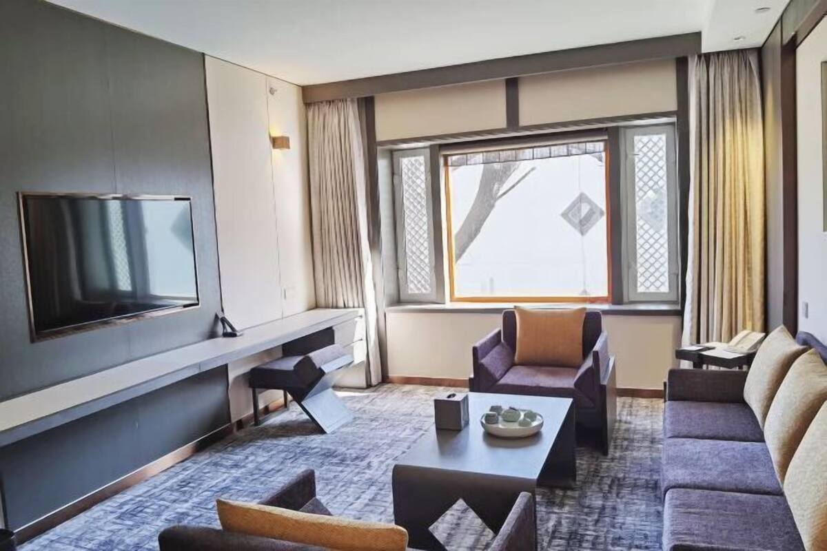 A modern hotel room with a flat-screen TV mounted on the wall, a sofa, a coffee table, and a large window with curtains.