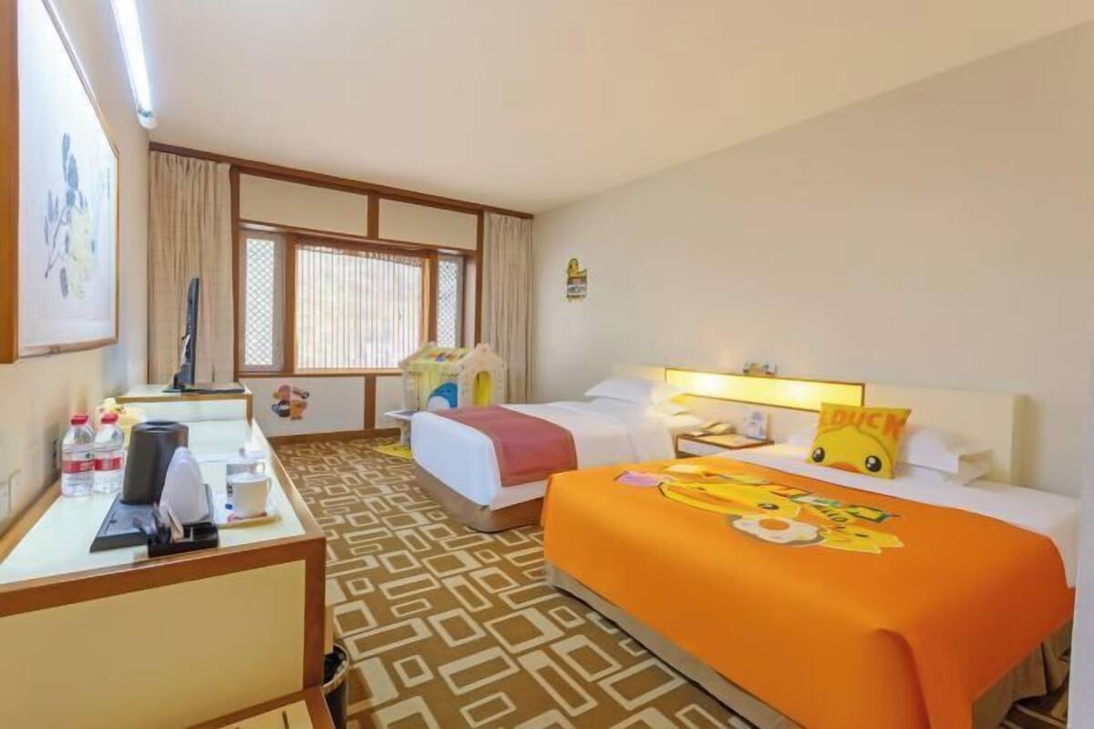 A hotel room with two beds, a desk with a chair, a window with curtains, and a wall-mounted light fixture.