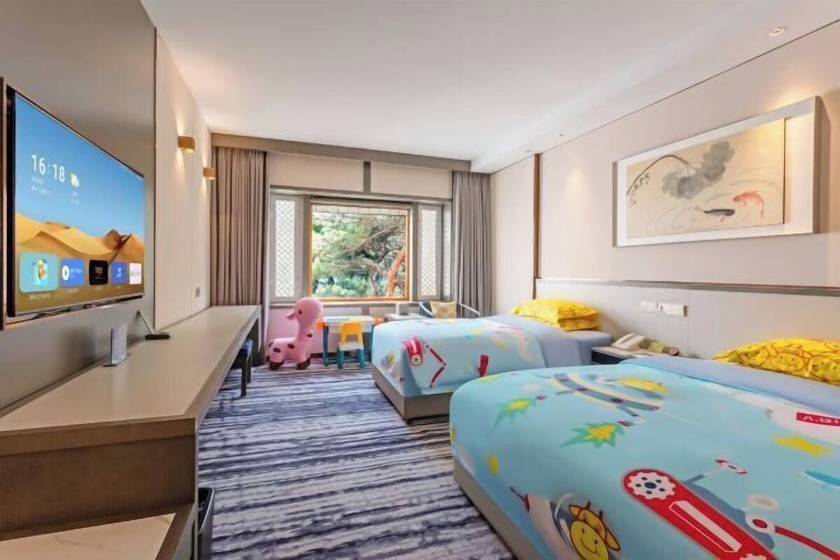 A hotel room with a TV, two beds with colorful bedding, a desk, a chair, a window with a view of greenery, and a painting on the wall.