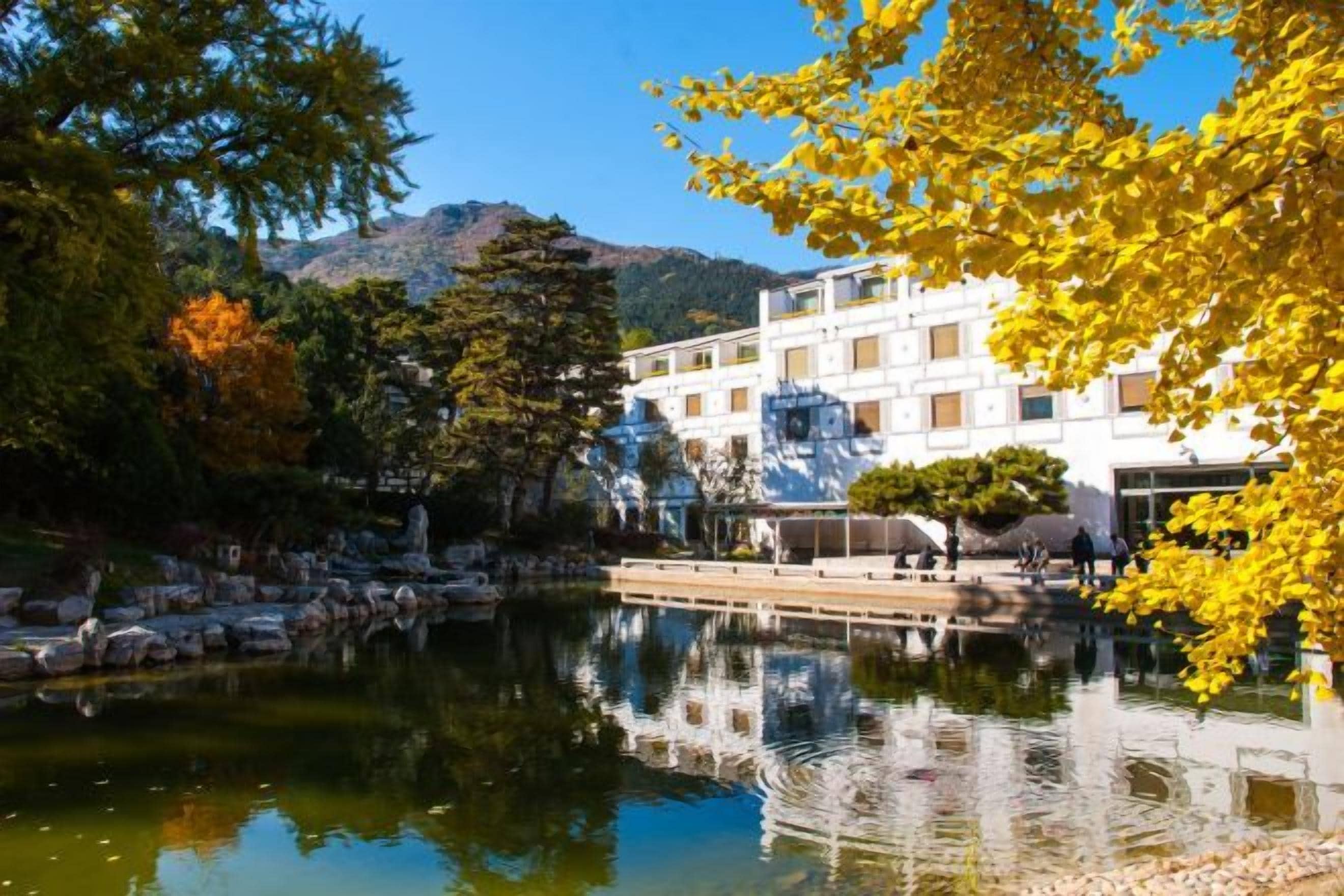 A building with a reflection in a calm body of water, surrounded by trees with autumn foliage.
