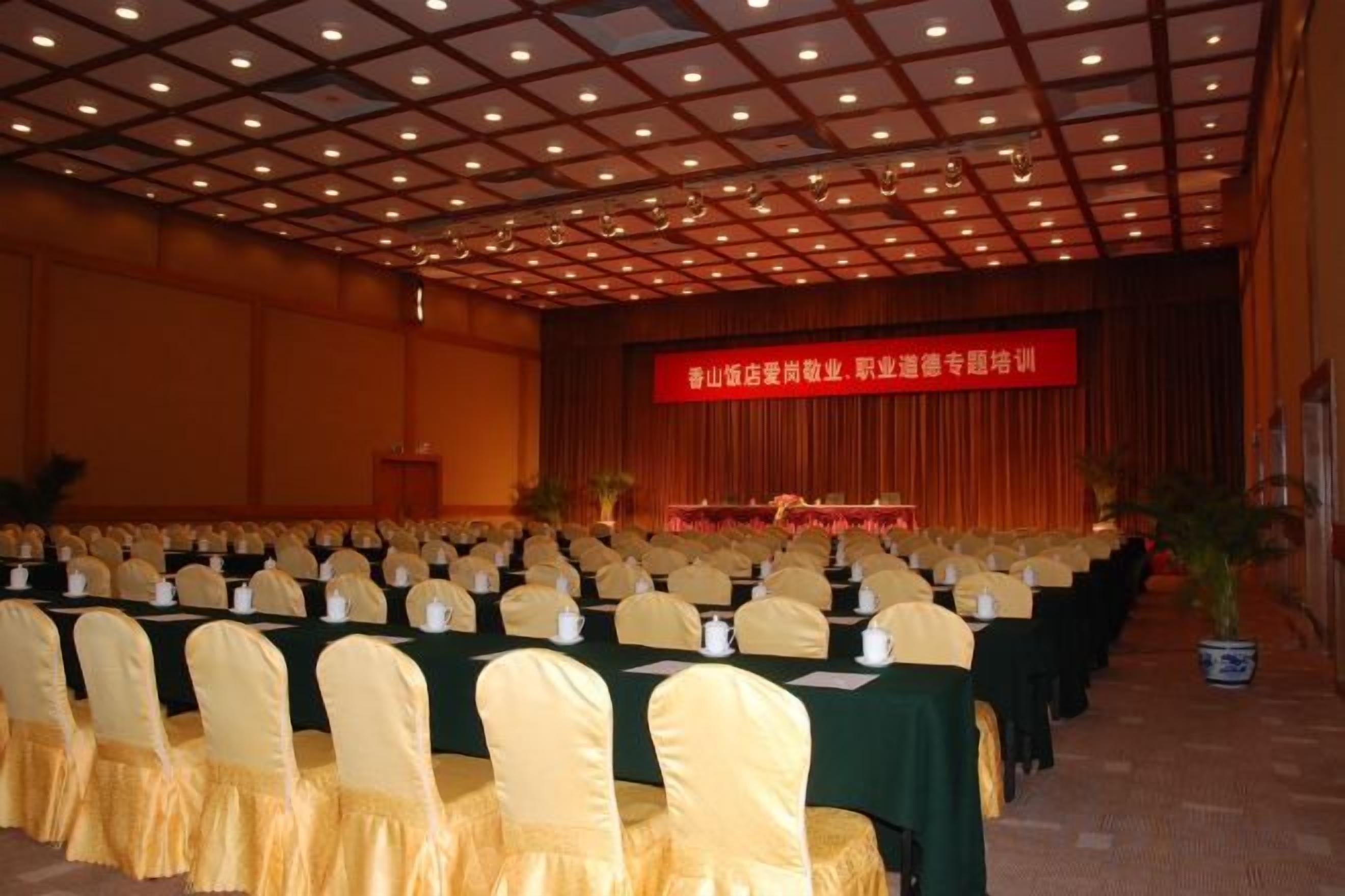 A conference room with rows of tables and chairs, a stage with a red banner, and a ceiling with a grid pattern.