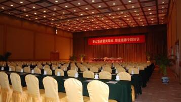 A conference room with rows of tables and chairs, a stage with a red banner, and a ceiling with a grid pattern.