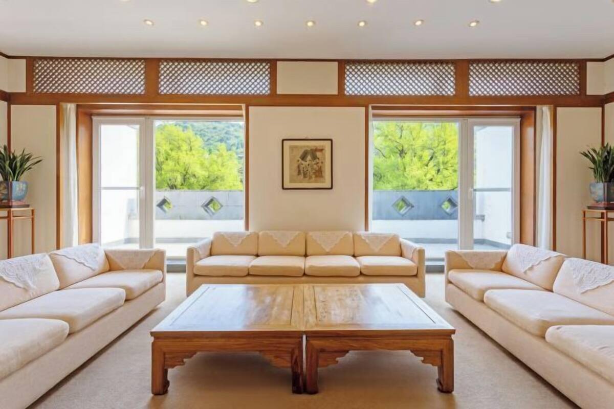 A spacious living room with a large wooden coffee table, two beige sofas, and a large window with a view of greenery.