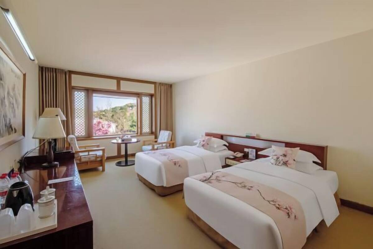 A hotel room with two beds, a desk, a chair, a lamp, and a window with a view of greenery.