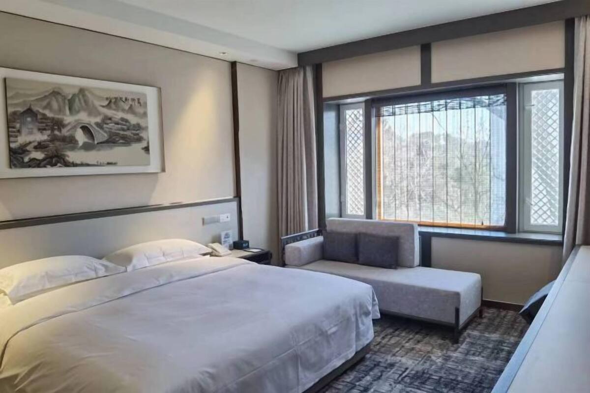 A hotel room with a bed, a bedside table, a chair, a window with a view, and a painting on the wall.