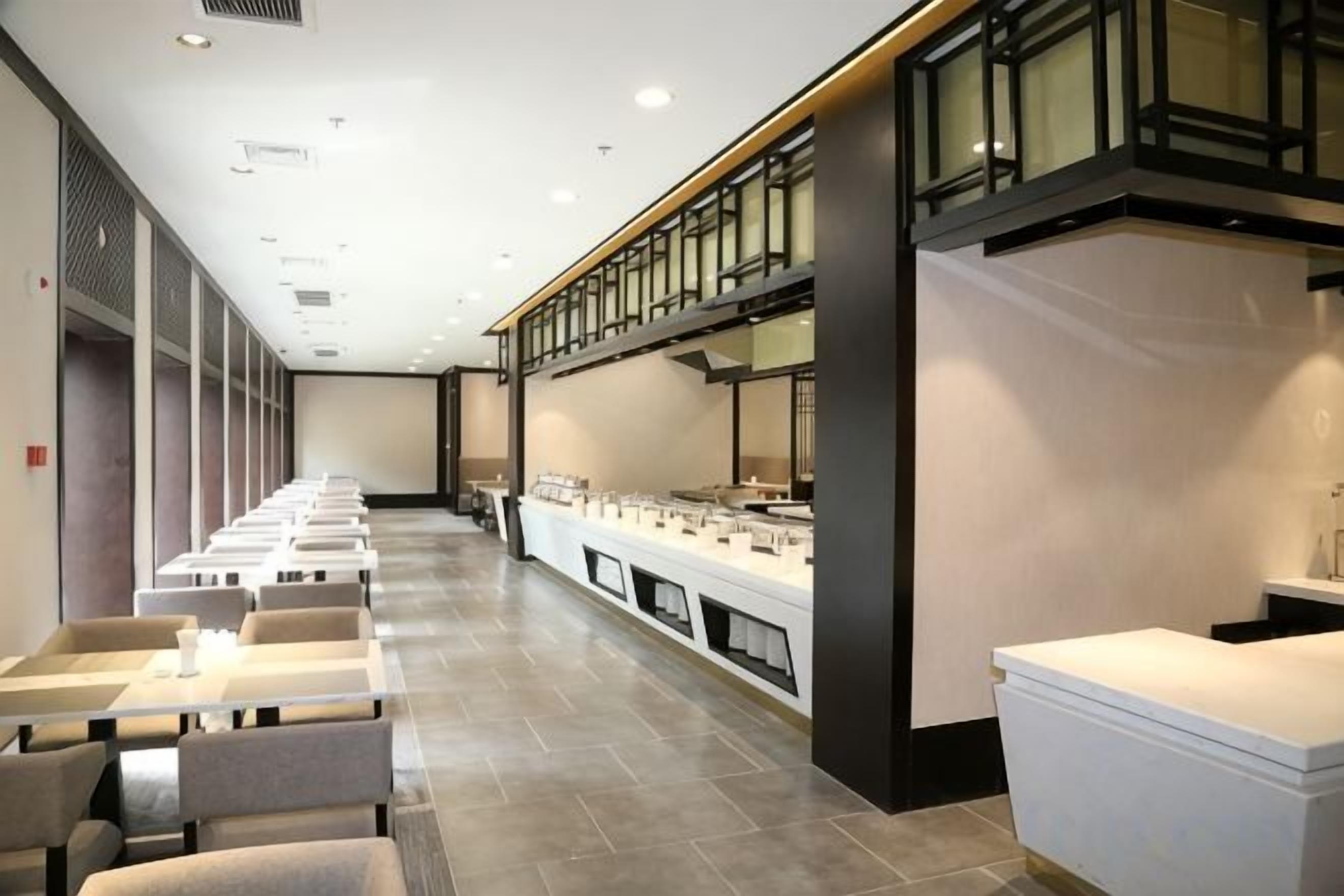 Una larga hilera de mesas y sillas en un moderno vestíbulo de hotel, con un mostrador y un espejo grande a la derecha.