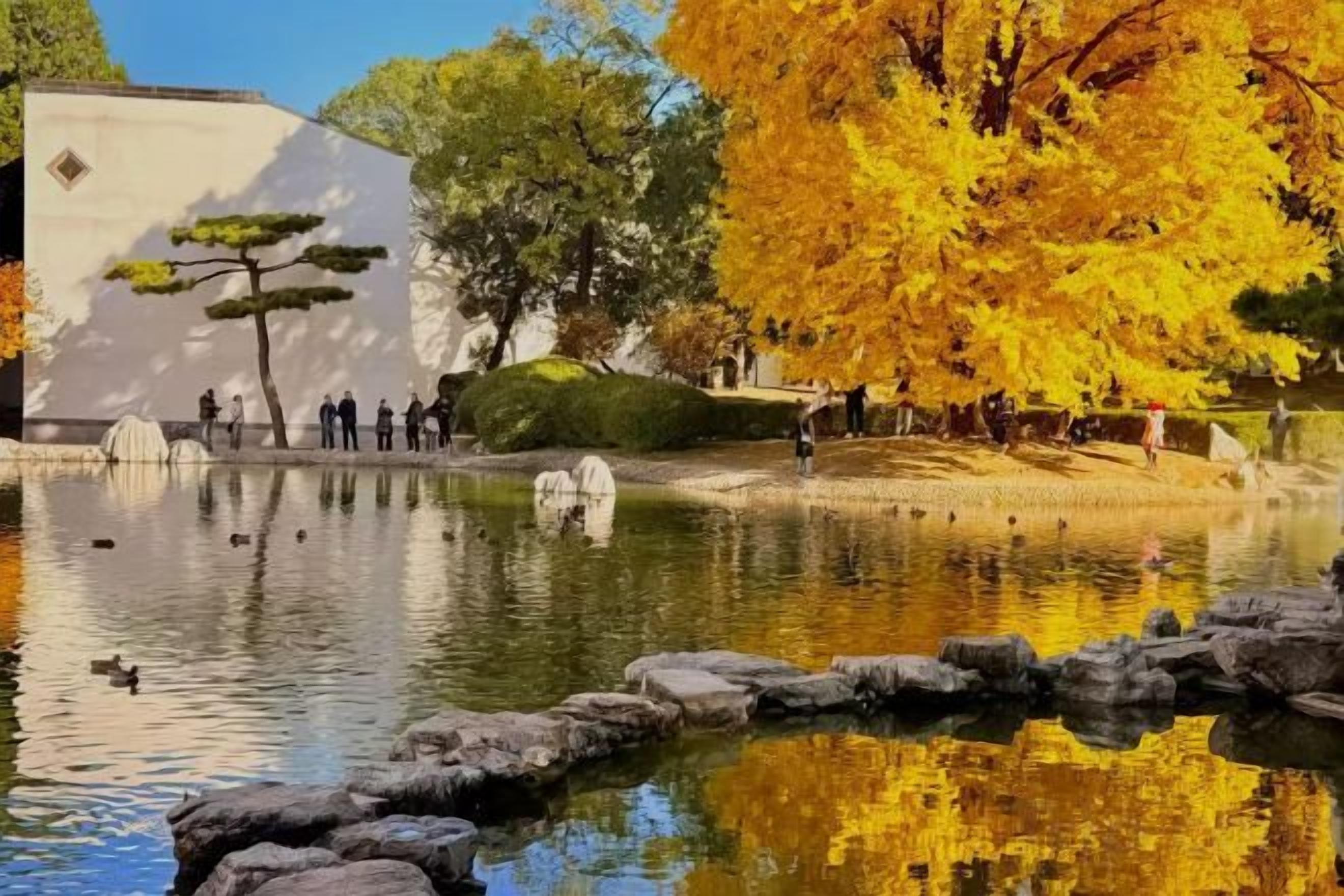 Un étang paisible, avec des rochers au premier plan, entouré d’arbres aux feuilles d’automne, et un bâtiment en arrière-plan.