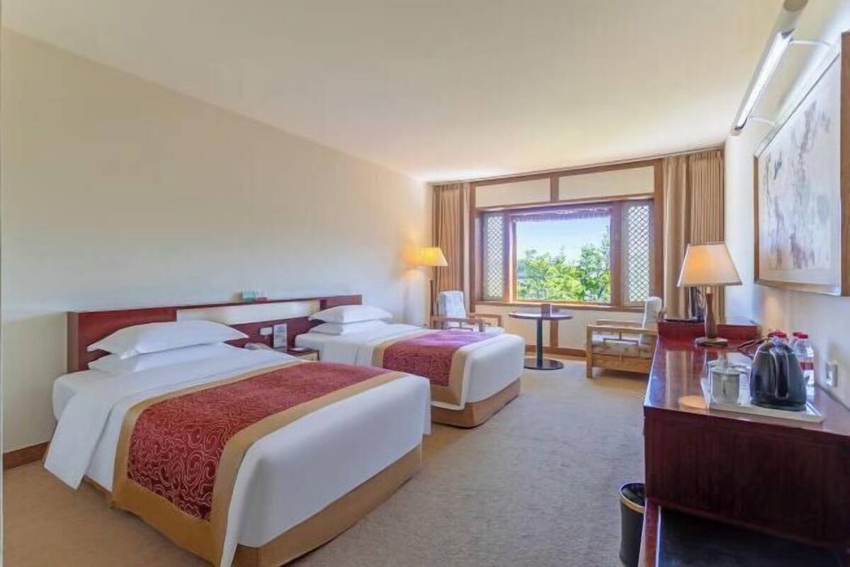 A hotel room with two beds, a desk, a chair, a window with a view, and a lamp.