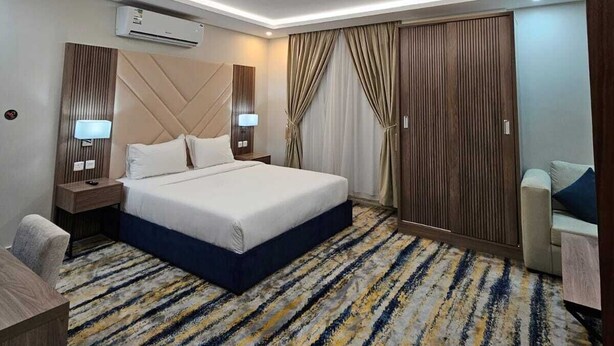 A modern hotel room with a large bed, a desk, a chair, and a sofa. There is an air conditioning unit on the wall and a ceiling light. - شقق أعمار الديار للشقق المخدومة  (Jeddah)