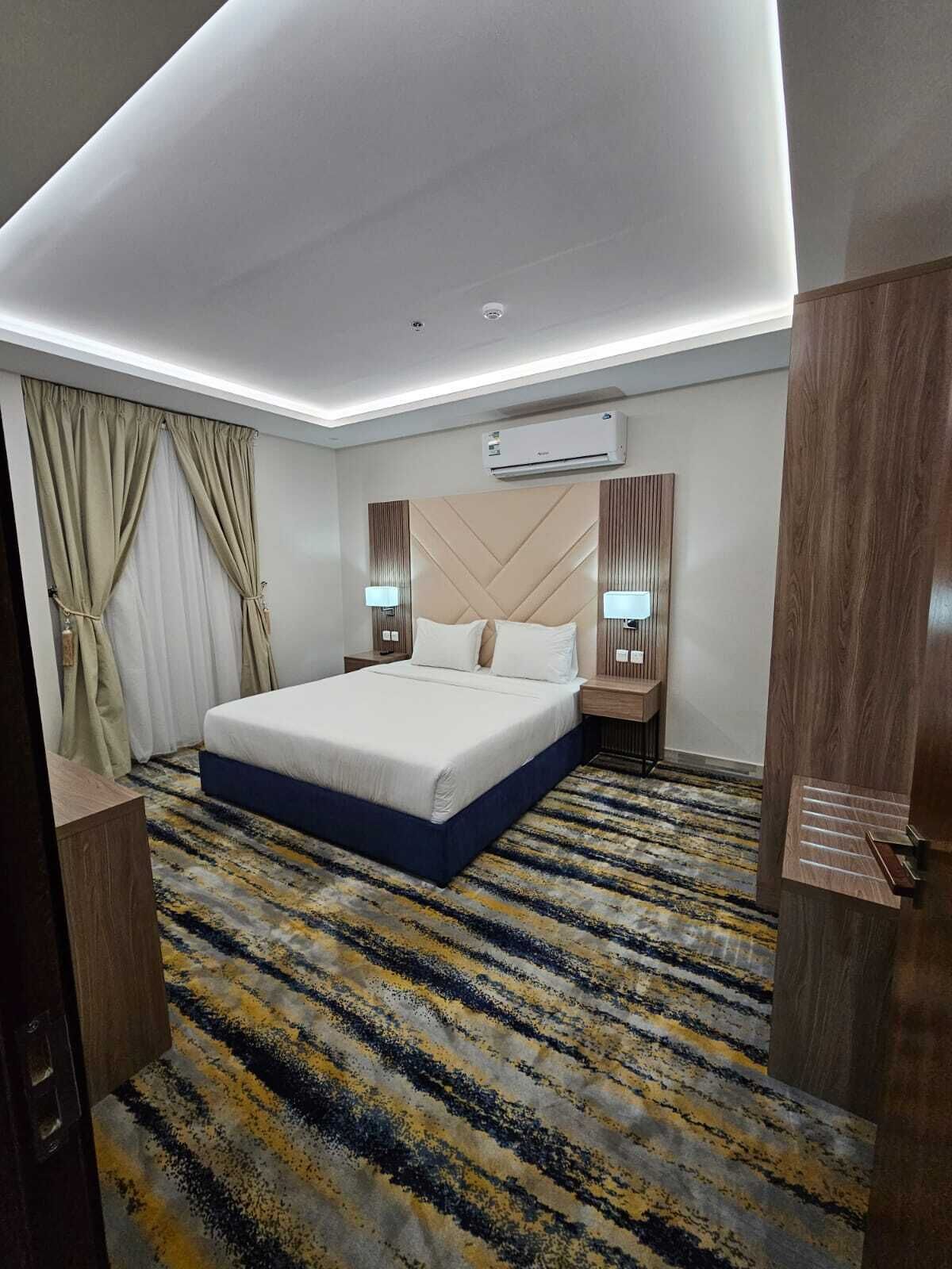 A hotel room with a large bed, bedside lamps, an air conditioning unit, and a patterned carpet.