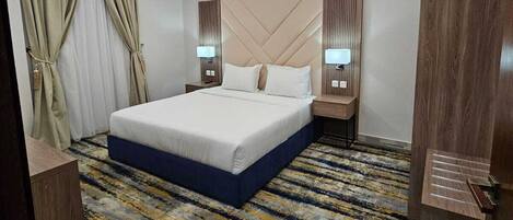 A hotel room with a large bed, bedside lamps, an air conditioning unit, and a patterned carpet.