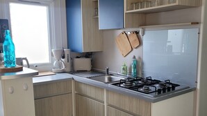 A compact kitchen with wooden cabinets, a gas stove, a coffee maker, and a window with a view. - Mobil Home 6 Personnes île D'oléron : Notre Oléronaise (Saint-Denis-d'Oléron)