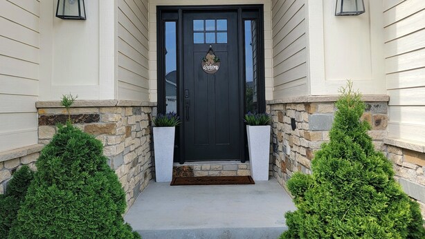 Une maison avec une porte d’entrée noire, une pancarte de bienvenue et deux plantes en pot de chaque côté de l’entrée.