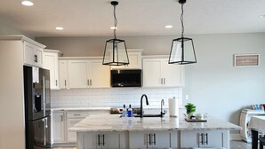 Une cuisine moderne avec des meubles blancs, un îlot central doté de tabourets de bar, un réfrigérateur en acier inoxydable et des luminaires suspendus.
