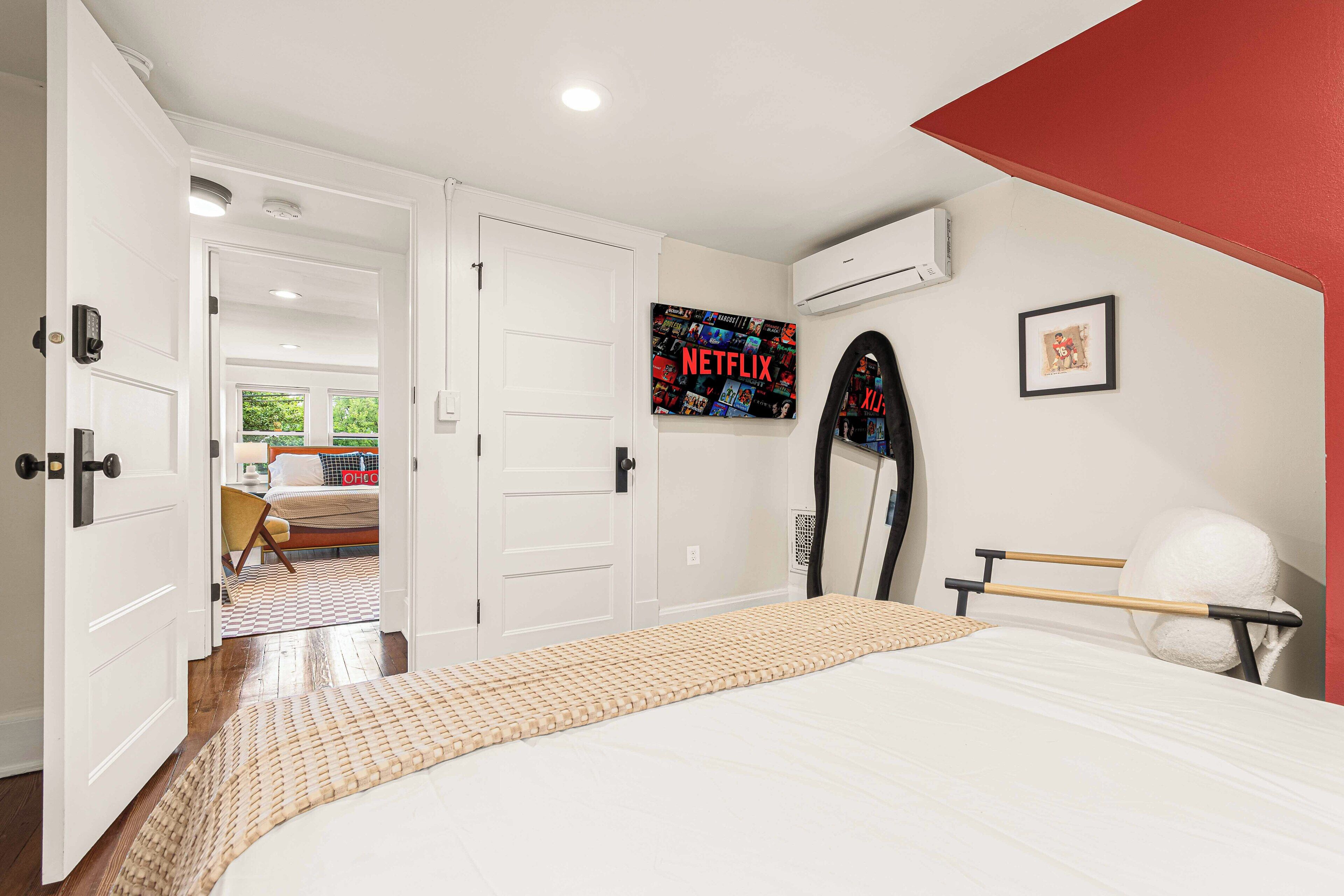 A bedroom with a bed, a wall-mounted air conditioner, a framed picture, and a door leading to another room.