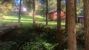 A wooden cabin surrounded by a lush forest, with a well-maintained lawn and a house in the background. - Tranquil cabin with WiFi in Brush Prairie (Brush Prairie)